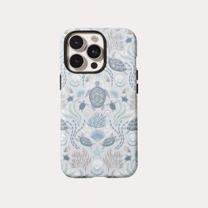 May include: A phone case with a sea-themed pattern. The design features turtles, seashells, coral, and seaweed in shades of blue and gray on a light gray background. The case has a black camera lens frame and black side buttons.
