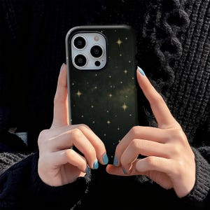 May include: A black phone case with a starry night design. The case features a dark background with numerous small, golden stars scattered across it. The phone case has a black border and cutouts for the phone's camera.