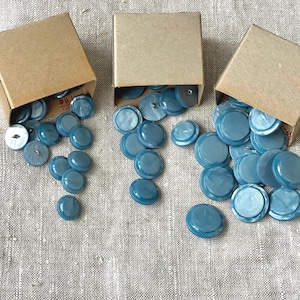 May include: Three boxes filled with light blue buttons. The boxes are made of cardboard and have a light brown color. The buttons are round and have a slightly iridescent finish.
