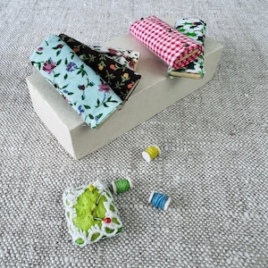 Puede incluir: Una colección de recortes de tela en varios patrones, incluyendo flores, cuadros y vichy, están dispuestos en una caja blanca. También hay cuatro carretes de hilo de diferentes colores y un alfiletero verde con ribete de encaje blanco y alfileres.