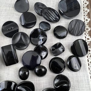 May include: A collection of various black buttons in different shapes and sizes. The buttons include round, oval, square, and triangular designs, some with textured surfaces. The buttons are arranged on a white, textured fabric.