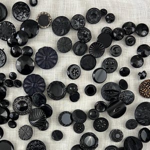 Könnte beinhalten: Eine Sammlung verschiedener schwarzer Knöpfe mit unterschiedlichen Formen und Texturen. Die Knöpfe variieren in der Größe und umfassen facettierte, florale und gemusterte Designs. Einige Knöpfe haben ein glänzendes Finish, andere sind matt.