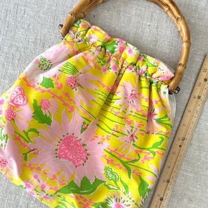 May include: A fabric handbag featuring a bright yellow and pink floral pattern with a curved bamboo handle. A wooden ruler is placed alongside the bag, showing a measurement of approximately 28 cm in height for the main body of the bag.