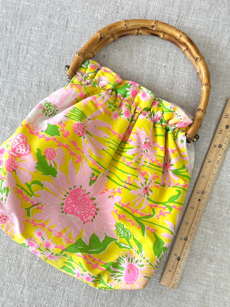 May include: A fabric handbag featuring a bright yellow and pink floral pattern with a curved bamboo handle. A wooden ruler is placed alongside the bag, showing a measurement of approximately 28 cm in height for the main body of the bag.