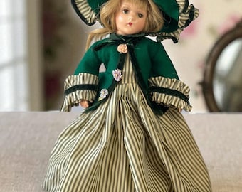 May include: A vintage doll wearing a green and white striped dress, a matching hat, and a green jacket with velvet trim. The doll's hat is adorned with pink and peach-colored flowers. The doll has blonde hair and is posed on a neutral surface.