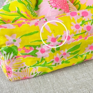 May include: A fabric bag featuring a vibrant floral pattern with bright yellow, pink, and green colours. The material is gathered at the top with a drawstring closure. A white circle highlights a small brown spot on the lower section of the fabric.