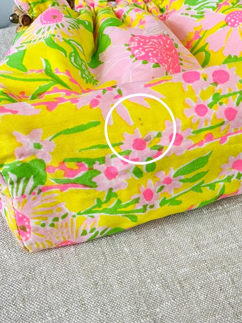 May include: A fabric bag featuring a vibrant floral pattern with bright yellow, pink, and green colours. The material is gathered at the top with a drawstring closure. A white circle highlights a small brown spot on the lower section of the fabric.
