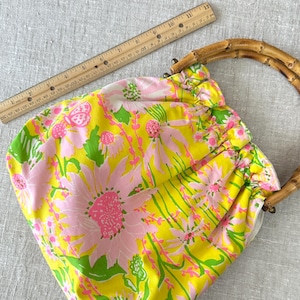 May include: A yellow fabric handbag featuring a pink and green floral pattern with a gathered top and two curved bamboo handles. A wooden ruler is positioned along the top edge, showing the bag width is approximately 28 cm across.