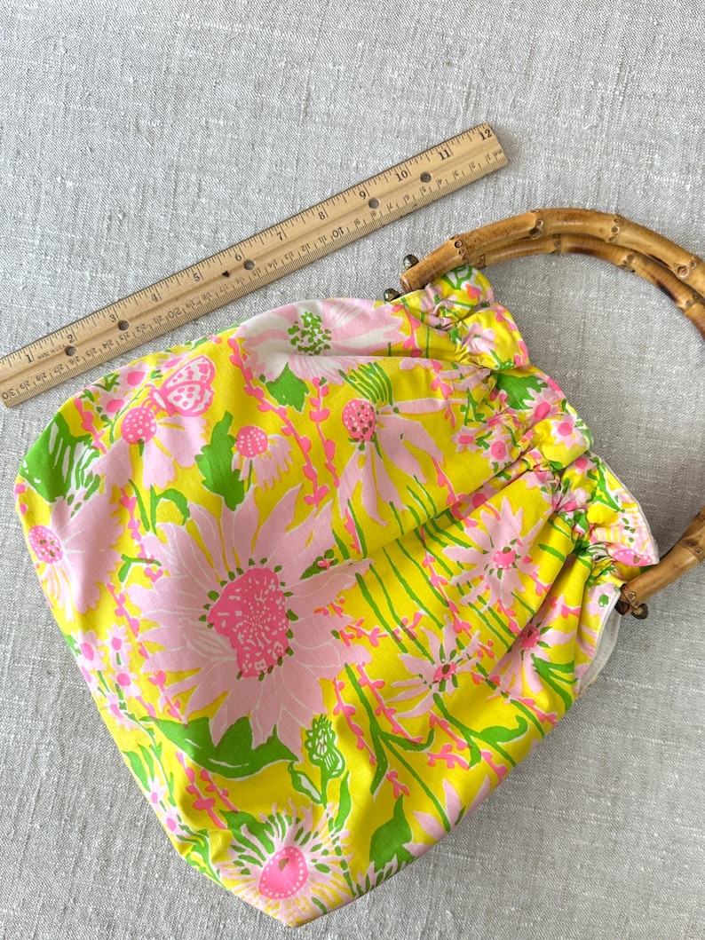 May include: A yellow fabric handbag featuring a pink and green floral pattern with a gathered top and two curved bamboo handles. A wooden ruler is positioned along the top edge, showing the bag width is approximately 28 cm across.