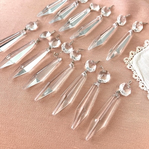 May include: Clear crystal chandelier prisms with faceted teardrop shapes and round crystal beads. The prisms are suspended from silver-toned metal pins, arranged on a pink surface. Decorative lighting components.