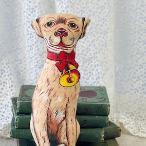 May include: A vintage stuffed dog doll with a tan body, red collar, and a yellow bell. The dog is sitting on a red base and has a detailed printed design. The doll is placed in front of a stack of antique green books.