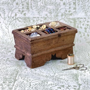 May include: A vintage wooden button box with multiple compartments filled with various buttons. A spool of thread and small scissors are next to the box. The box is brown and sits on a patterned surface.