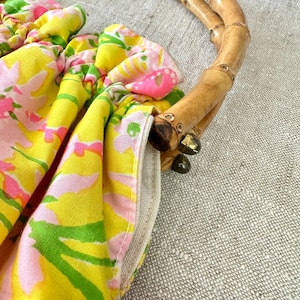 May include: A handbag featuring a bright yellow fabric with a pink and green floral pattern, gathered at the top. The bag is equipped with two curved, natural bamboo handles attached to the fabric with small, dark metal hardware components.
