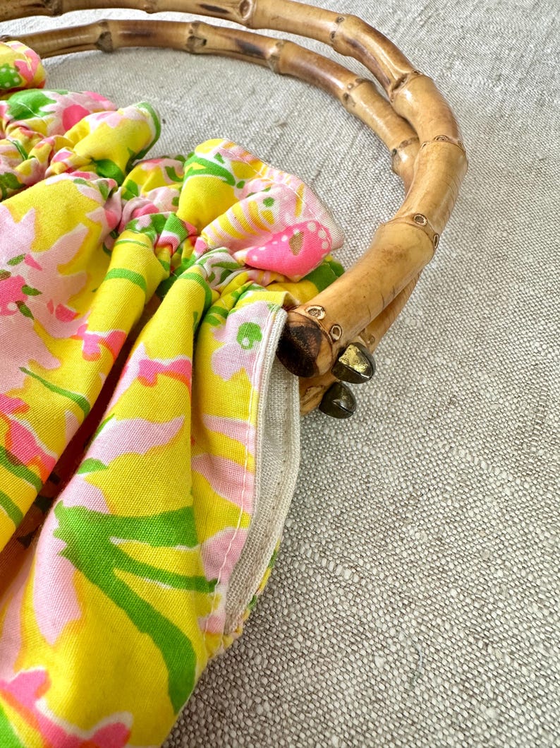 May include: A handbag featuring a bright yellow fabric with a pink and green floral pattern, gathered at the top. The bag is equipped with two curved, natural bamboo handles attached to the fabric with small, dark metal hardware components.