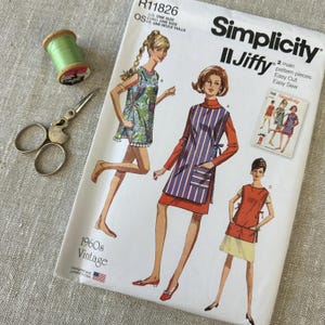 May include: A Simplicity Jiffy sewing pattern envelope R11826 for a 1960s vintage garment, featuring illustrations of three variations of a sleeveless tunic or apron. Includes a spool of light green thread and a pair of metal sewing scissors.