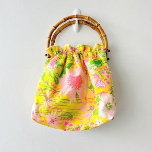 May include: A bright yellow fabric handbag featuring a vibrant floral pattern in shades of pink, white, and green. The bag has a gathered, ruffled top edge and two rounded, natural bamboo handles that provide a sturdy grip for carrying the item.