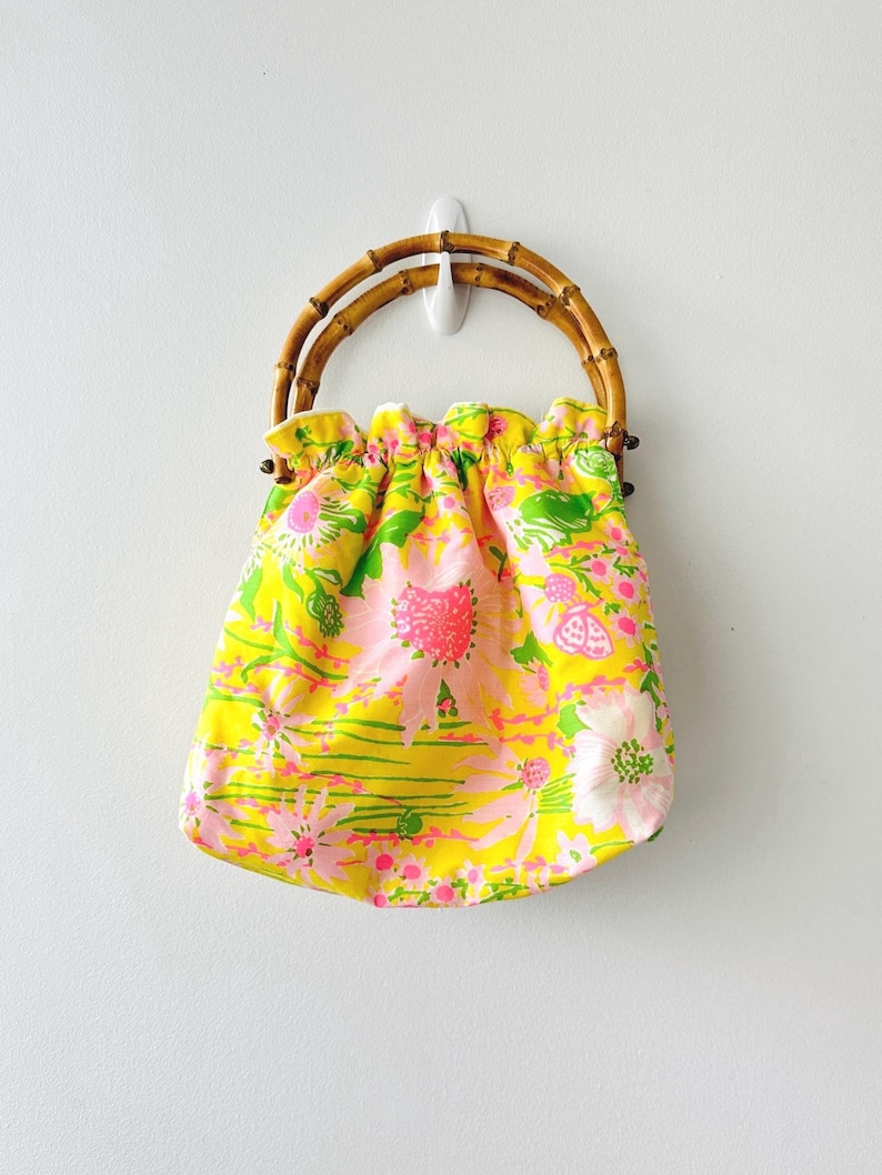 May include: A bright yellow fabric handbag featuring a vibrant floral pattern in shades of pink, white, and green. The bag has a gathered, ruffled top edge and two rounded, natural bamboo handles that provide a sturdy grip for carrying the item.