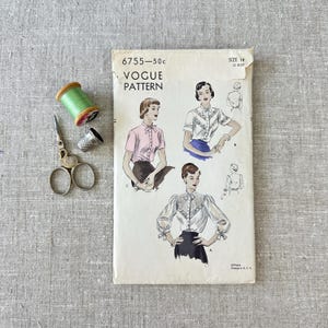May include: A vintage Vogue sewing pattern envelope numbered 6755, size 14, featuring illustrations of three blouse variations. Beside the envelope are a spool of light green thread and a small silver-colored metal sewing thimble.