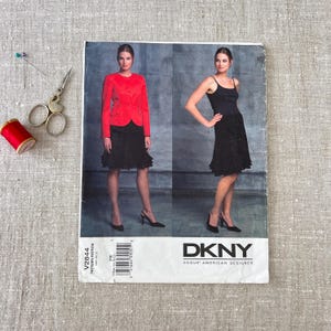 May include: A sewing pattern for a DKNY Vogue American Designer dress. The pattern features a woman in a red jacket and black skirt, and another in a black dress. A spool of red thread, scissors, and pins are on the left.