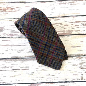 May include: A patterned tie in shades of brown, blue, and purple, with a classic plaid design. The tie is made of a textured fabric and is displayed on a weathered wooden surface. The tie is a classic accessory.