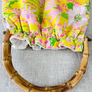 May include: A fabric bag featuring a vibrant floral pattern in shades of yellow, pink, and green. The bag has a gathered top opening and a rounded handle made of natural bamboo with visible segments and a light brown finish.