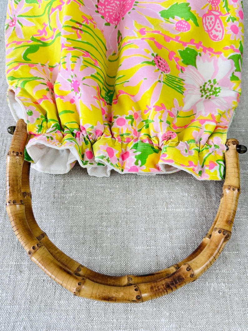 May include: A fabric bag featuring a vibrant floral pattern in shades of yellow, pink, and green. The bag has a gathered top opening and a rounded handle made of natural bamboo with visible segments and a light brown finish.