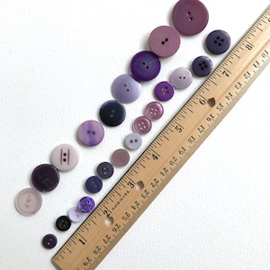 May include: A collection of purple buttons of varying sizes and shades arranged in a line on a white surface. A wooden ruler with inch markings is placed diagonally across the buttons.