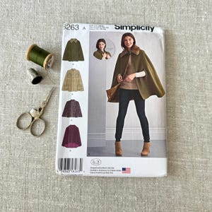 May include: A Simplicity sewing pattern envelope 8263 featuring illustrations of capes in olive, tan, tweed, and plum, alongside a spool of olive green thread, a metal thimble, and silver sewing scissors with ornate handles on a textured fabric.