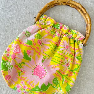 May include: A fabric handbag featuring a bright yellow background with a repeating pattern of pink flowers, green leaves, and small butterflies. The bag has a gathered top opening and a curved, segmented bamboo handle attached with metal hardware.