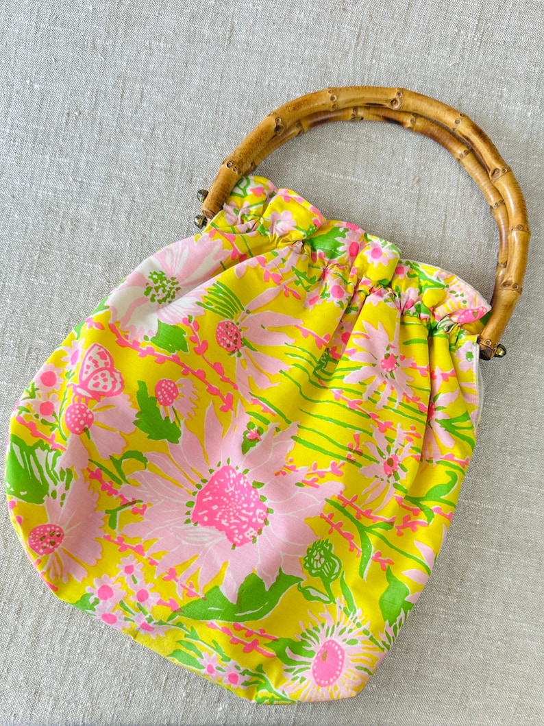 May include: A fabric handbag featuring a bright yellow background with a repeating pattern of pink flowers, green leaves, and small butterflies. The bag has a gathered top opening and a curved, segmented bamboo handle attached with metal hardware.