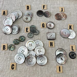 Peut inclure: Assortiment de boutons en nacre de différentes tailles et formes, étiquetés de A à L. Les boutons sont disposés sur un fond en tissu brun clair.