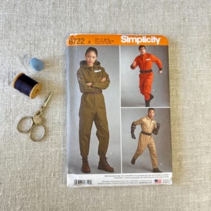 Könnte beinhalten: Ein Simplicity 8722 Schnittmusterumschlag mit Abbildungen von Overalls in Olivgrün, Orange und Beige. Der Umschlag zeigt den Markennamen, die Modellnummer und die Größen XS-XL, begleitet von einem blauen Fingerhut, schwarzem Garn und einer Schere.