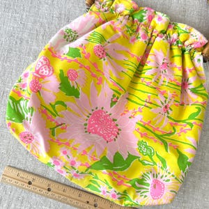 May include: A fabric pouch bag with a bright yellow background and a floral pattern in pink and green, featuring a rounded bamboo handle. A wooden ruler is placed along the bottom edge, showing the bag width is approximately 30.5 cm.