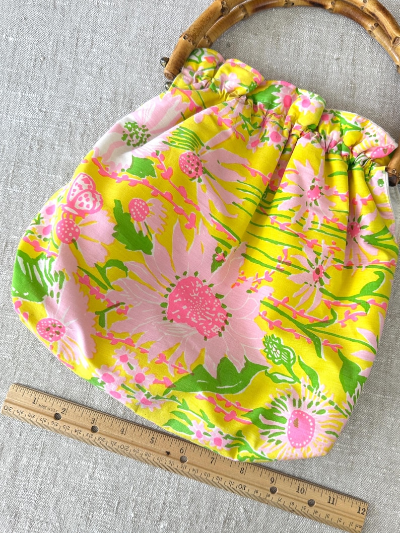 May include: A fabric pouch bag with a bright yellow background and a floral pattern in pink and green, featuring a rounded bamboo handle. A wooden ruler is placed along the bottom edge, showing the bag width is approximately 30.5 cm.