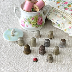 Op de afbeelding: Een verzameling vintage naaibenodigdheden, waaronder verschillende metalen vingerhoeden, een suikerpot met bloemmotief gevuld met roze garenklosjes en een kleine bloemenpin. De items zijn gerangschikt op een getextureerd, neutraal gekleurd oppervlak.