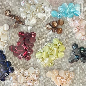 May include: A collection of round buttons in various colors including white, light blue, brown, dark red, yellow, black, and beige, each set packaged in individual clear plastic bags, showcasing a smooth finish and central holes.