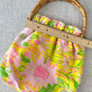 May include: A fabric handbag featuring a bright yellow background with a pink and green floral pattern and a curved bamboo handle. A wooden ruler is placed across the top opening, showing a width measurement of approximately 23 centimetres.