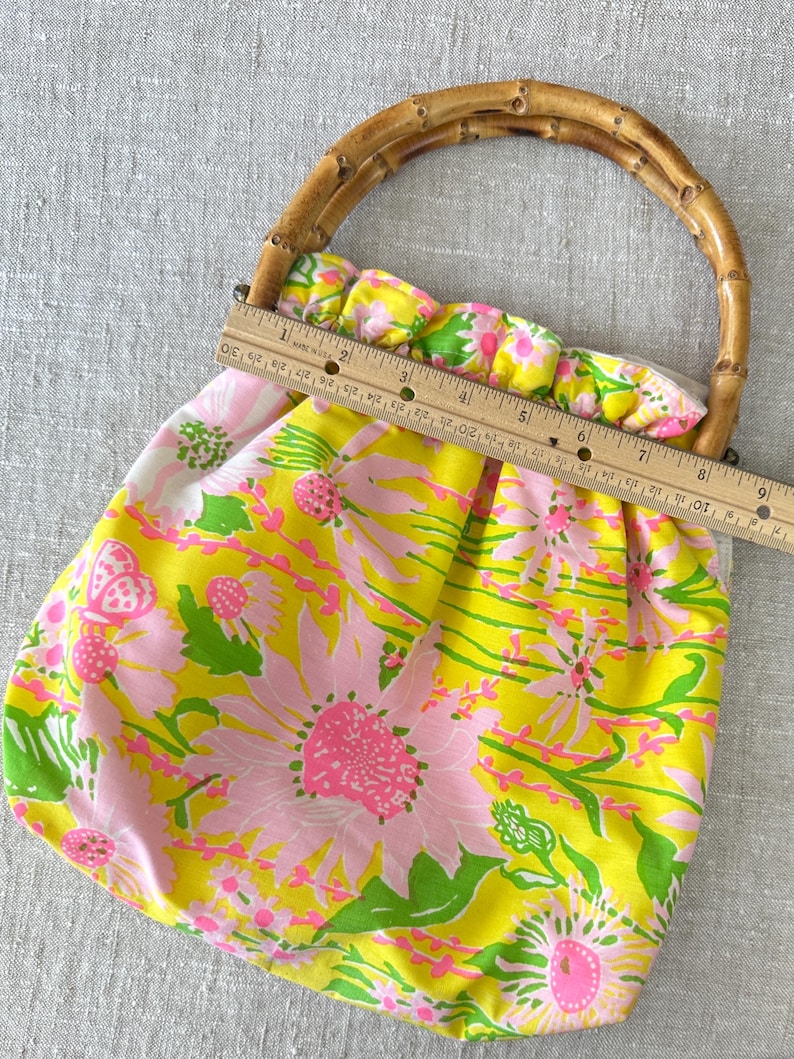 May include: A fabric handbag featuring a bright yellow background with a pink and green floral pattern and a curved bamboo handle. A wooden ruler is placed across the top opening, showing a width measurement of approximately 23 centimetres.