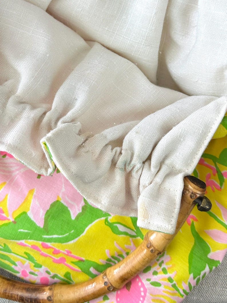 May include: A handbag featuring a bright yellow fabric exterior with a pink and green floral pattern. The bag has a gathered off-white linen lining and a curved, segmented bamboo handle attached to the fabric with stitched fabric loops.