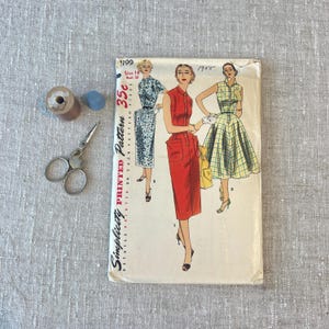 May include: A vintage Simplicity printed sewing pattern with dress designs. The pattern packet shows illustrations of dresses in red, blue, and yellow. Also included are a wooden spool, blue thimble, and small scissors.