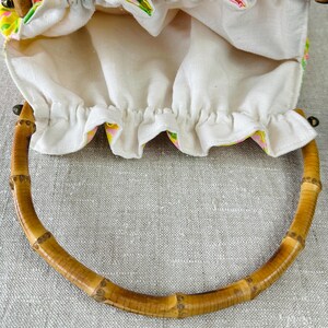 May include: A drawstring pouch bag featuring a vibrant floral pattern in shades of pink, yellow, and green. The bag has a gathered white fabric top and two curved, segmented bamboo handles attached to the sides with small metal hardware.