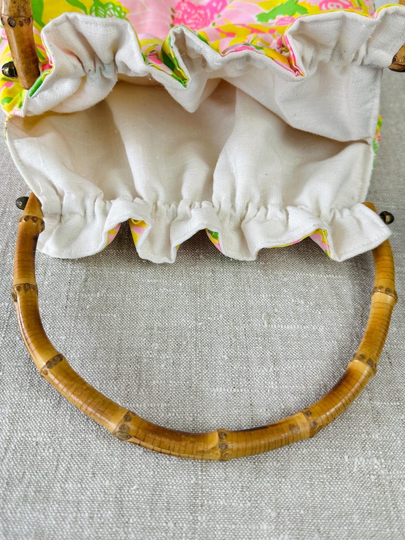 May include: A drawstring pouch bag featuring a vibrant floral pattern in shades of pink, yellow, and green. The bag has a gathered white fabric top and two curved, segmented bamboo handles attached to the sides with small metal hardware.