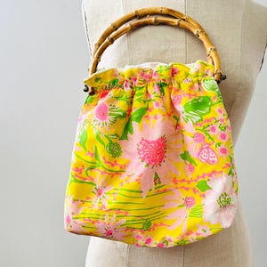 May include: A drawstring handbag featuring a bright yellow fabric with a vibrant floral pattern in shades of pink, white, and green. The bag is equipped with a rounded, natural bamboo handle attached to the top, creating a gathered, soft silhouette.