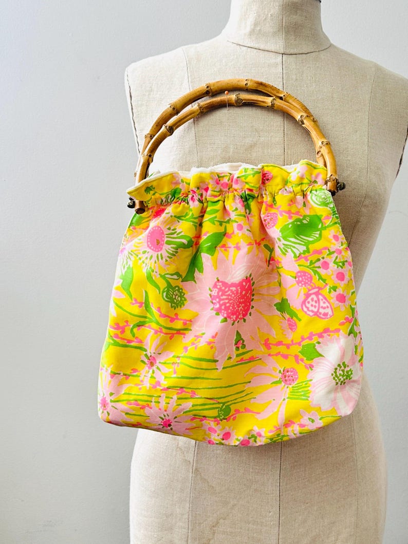 May include: A drawstring handbag featuring a bright yellow fabric with a vibrant floral pattern in shades of pink, white, and green. The bag is equipped with a rounded, natural bamboo handle attached to the top, creating a gathered, soft silhouette.