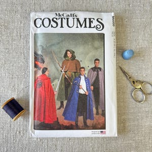 May include: A McCall's Costumes sewing pattern envelope M6335 featuring illustrations of three capes in red, green, and blue. The white envelope includes text, a spool of dark blue thread, a blue thimble, and metal sewing scissors.