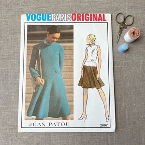 May include: A vintage Vogue Paris Original sewing pattern featuring two designs by Jean Patou. The pattern is for a dress and a skirt. The dress is blue and the skirt is brown. The pattern number is 2697.