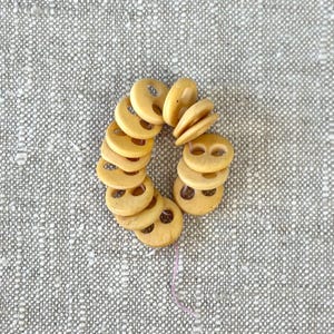 May include: A circular arrangement of light tan, two-hole buttons strung together on a thin, light-colored thread. Each button features a smooth, rounded surface with a consistent pale yellow-brown hue and two central holes for attachment.