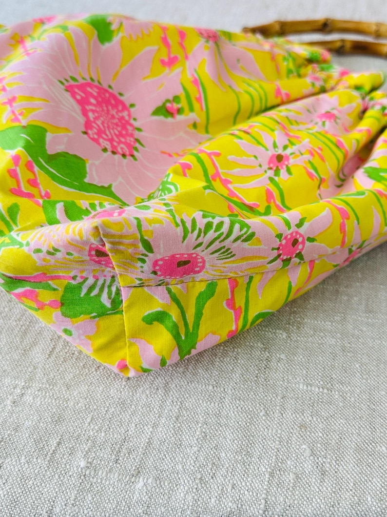 May include: A fabric handbag featuring a vibrant floral pattern with large pink and white flowers, bright green leaves, and a yellow background. The bag includes a curved bamboo handle attached to the top, showcasing a gathered, soft construction.