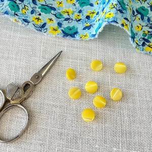 May include: A collection of yellow, round buttons with a white stripe, scattered on a textured, light gray surface. A pair of silver scissors and a piece of floral fabric with blue and yellow flowers are also present.