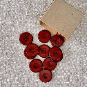 Könnte beinhalten: Eine Gruppe von 10 roten Holz-Knöpfen mit jeweils zwei Löchern. Die Knöpfe sind in einer Halbkreisform angeordnet und liegen auf einer hellbraunen Stoffoberfläche.
